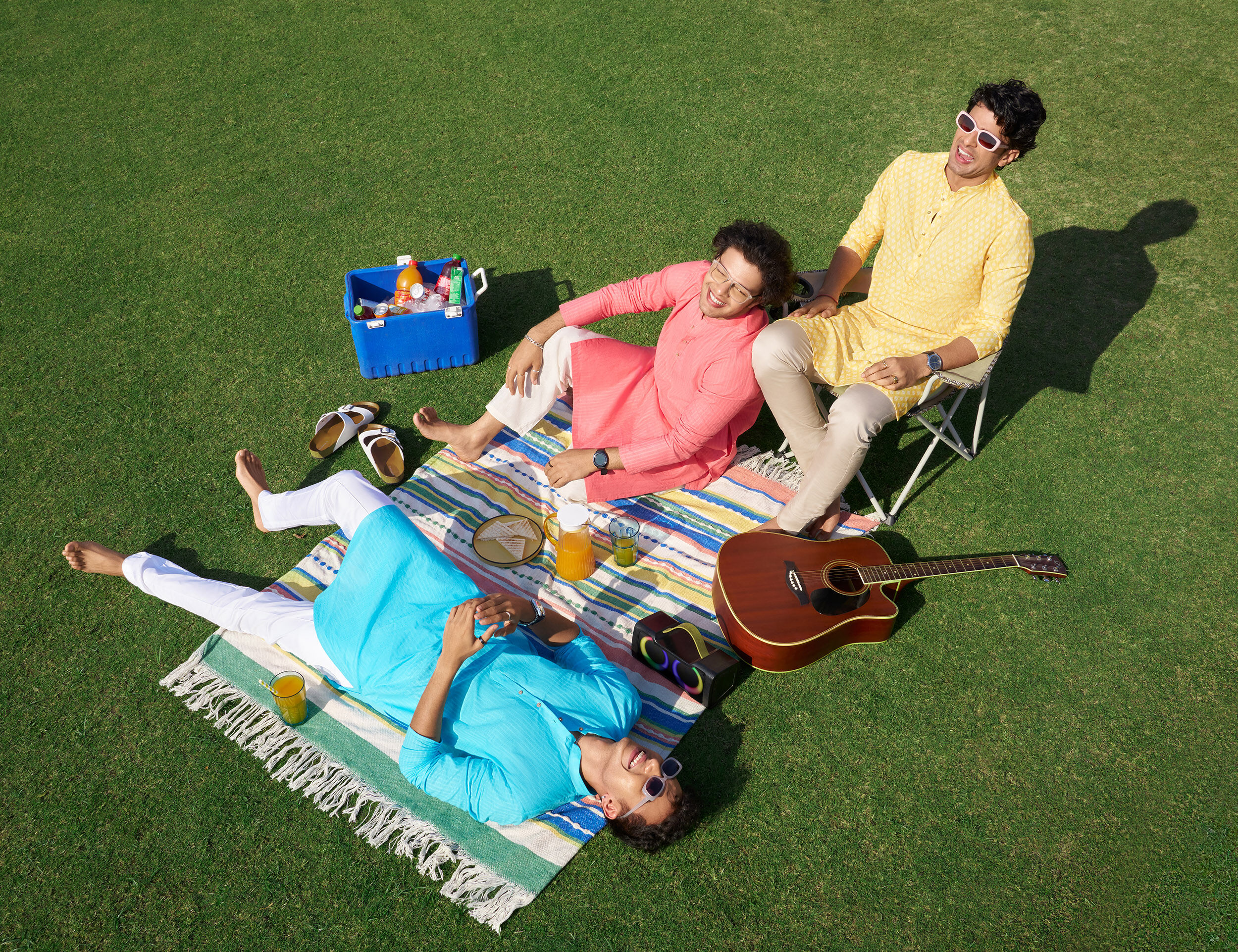 Men in vibrant cotton kurtas relaxing outdoors on a sunny day, enjoying a summer picnic with drinks and a guitar.